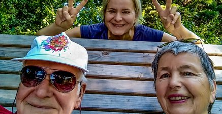 Älteres Paar auf einer hölzernen Liegebank, dahinter eine jüngere Frau in Sportkleidung.