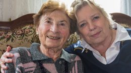 Eine alte Frau und eine Frau mittleren Alters sitzen nebeneinander auf einem Sofa. Die Jüngere von beiden hat ihren Arm um die alte Frau gelegt. Sie hält den Kopf zu ihr geneigt.