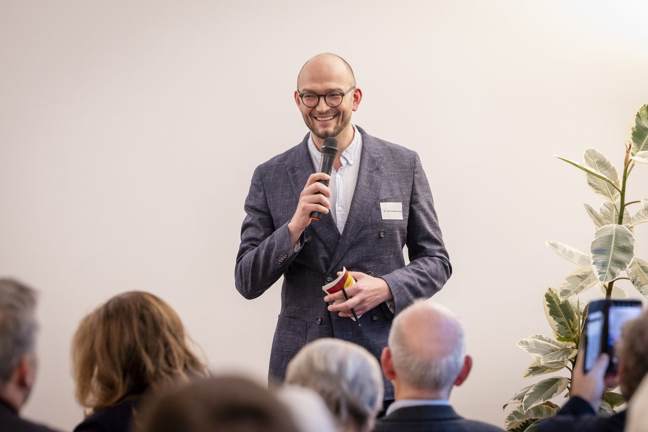 Ein junger Mann steht vor einem Publikum und spricht in ein Mikrofon. In der anderen Hand hält er Karteikarten und einen Stift. Er trägt ein dunkles Jackett und darunter ein helles Hemd. Klick öffnet eine vergrößerte Ansicht.