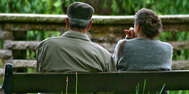 Ein Mann und eine Frau mit grauen Haaren sitzen auf einer Bank im Grünen. Sie sitzen mit dem Rücken zur Kamera.