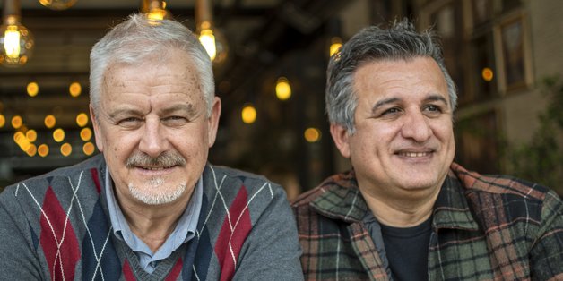 Zwei ältere Männer sitzen nebeneinander und schauen nach vorne. Einer hat graue Haare, trägt einer grauen Pullover mit kariertem Muster. Der ander hat dunkle, graumelierte Haare. Er trägt eine karierte Jacke, darunter ein schwarzes Shirt.