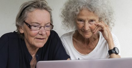 Zwei ältere Frauen bicken aufmerksam auf den Bildschirm eines Laptops. 