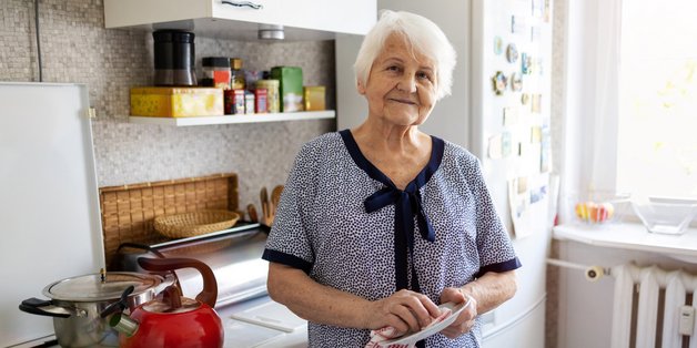 Alte Frau steht in der Küche vor ihrem Herd. Sie hält ein Geschirrtuch in der Hand.