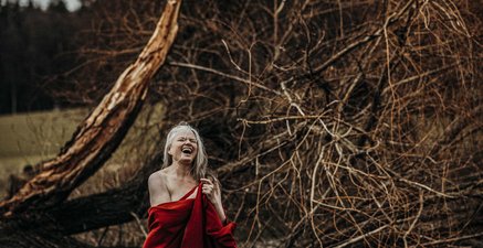 Herzlich lachende Frau mit weißem Haar steht in ein rotes Tuch gehüllt mit nackten Füßen in der Natur
