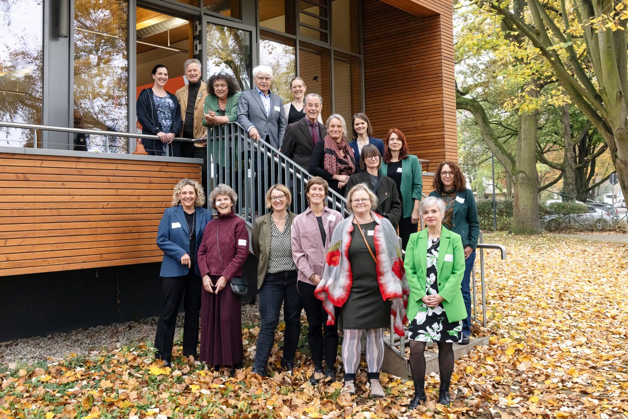 Eine Personengruppe Gruppe mit Menschen unterschiedlichen Alters steht vor und auf einer Treppe für ein Gruppenfoto. Im Hintergrund ist das Veranstaltungsgebäude mit einer Holz- und Glasfassade zu sehen.