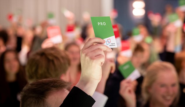 Viele sitzende Menschen in einem Raum. Sie haben Karten in ihren Händen und halten sie nach oben. Im Vordergrund sieht man eine Hand, mit einer grünen Karte. Daauf steht das Wort PRO. Klick öffnet eine vergrößerte Ansicht.