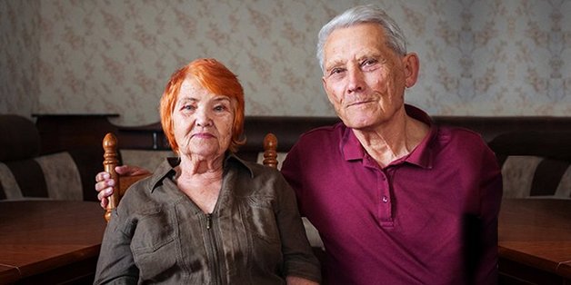 Ein älterer Mann und eine ältere Frau sitzen nebeneinander in einem Wohnzimmer. Der Mann hat den Arm um die Schulter der Frau gelegt.
