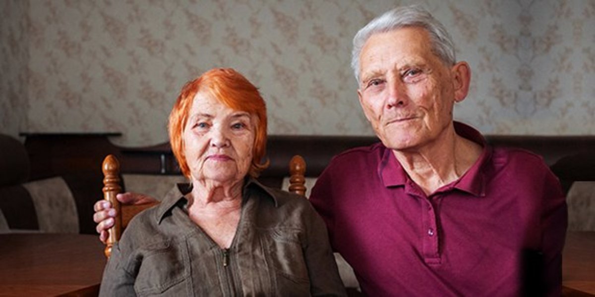 Ein älterer Mann und eine ältere Frau sitzen nebeneinander in einem Wohnzimmer. Der Mann hat den Arm um die Schulter der Frau gelegt.