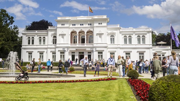 Blick auf die Villa Hammerschmidt in Bonn. Ein neoklassisischtisches Gebäude. Davor ein Blick auf dem Park mit Besucherinnen und Besuchern.