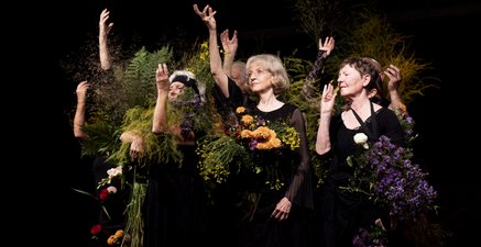 ältere Tänzer*innen stehen dem Licht zugeneigt mit bunten Gräsern & Blumen in einem Pulk zusammen