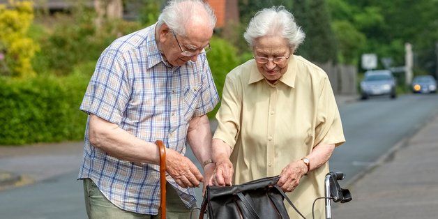 Ein alter Mann mit Gehstock und eine alte Frau mit Rollator stehen auf dem Bürgersteig. Sie schauen in eine Einkaufstasche, die auf dem Rollator abgestellt ist.