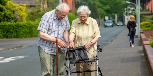 Ein alter Mann mit Gehstock und eine alte Frau mit Rollator stehen auf dem Bürgersteig. Sie schauen in eine Einkaufstasche, die auf dem Rollator abgestellt ist.