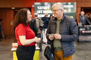 Eine Frau mittleren Alters und ein älter Mann unterhalten sich. Sie hat krause dunkle, schulterlange Haare und hälte eine Stapel kleiner Karten in der Hand. Der Mann hat graue, kurze Haare. Trägt Pullover und Cordhose. Ich den Händen hält er eine Karte.