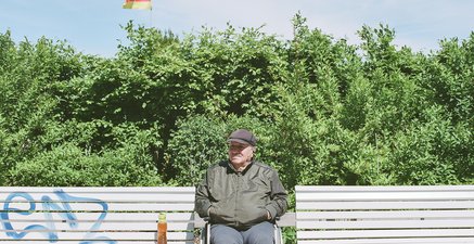 Älterer Mann mit Kappe im Rollstuhl zwischen zwei Parkbänken schaut in die Ferne. Die Sonne scheint. 