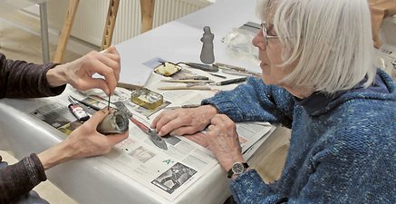 Ältere Frau sitzt an einem Tisch und beobachtet eine Person bei kunsthandwerklicher Tatigkeit