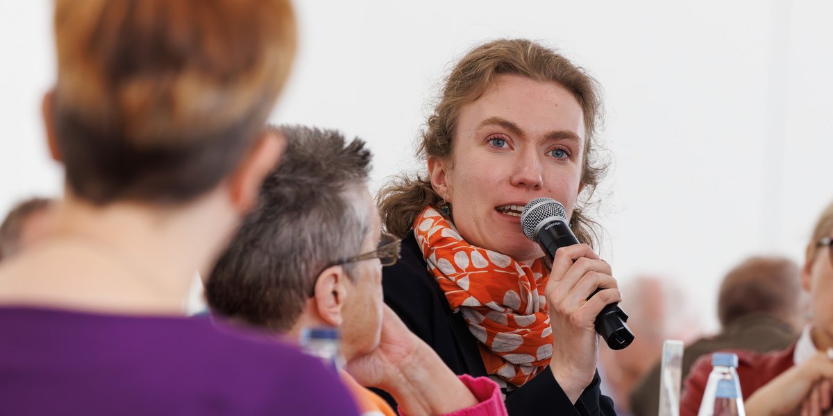 Ein Veranstaltungsraum. An Tischen sitzen ältere und junge Menschen. Ein junge Frau spicht gerade in ein Mikrofon, die anderen hören aufmerksam zu.