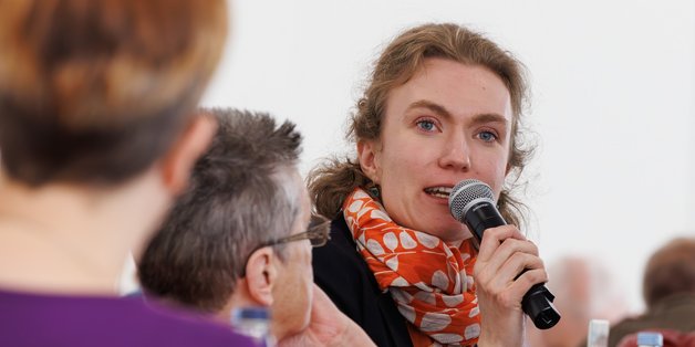 Ein Veranstaltungsraum. An Tischen sitzen ältere und junge Menschen. Ein junge Frau spicht gerade in ein Mikrofon, die anderen hören aufmerksam zu.