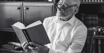 Älterer Mann liest Buch mit zwei Brillen auf der Nase. Im Hintergrund ein Bücherregal.