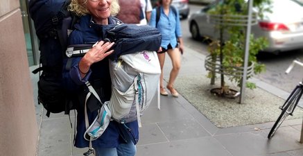 Ältere Frau steht mit Rucksack vorne und Rucksack hinten auf einem Bürgersteig