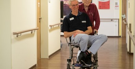 Mitarbeiter Ü60 wird von einer Seniorin Ü80 auf ihrem Rollator über den Flur geschoben.