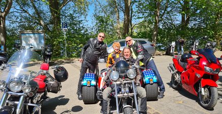 Ein Foto mit Motorrädern. In der Mitte ein Trike, mit drei Männern und einer älteren Frau