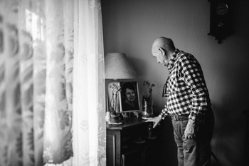 Links eine Gardine, rechts schaut ein älterer Mann auf ein gerahmtes Foto seiner Frau.