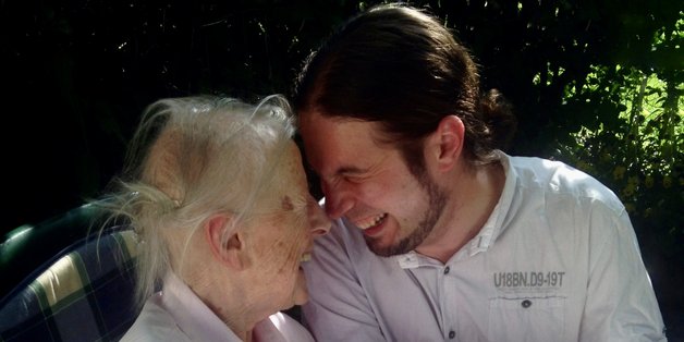 Eine alte Frau im Rollstuhle sitzt mit ihrem Enkel im Garten. Sie stecken die Köpfe zusammen und lachen gemeinsam.