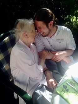 Eine alte Frau im Rollstuhle sitzt mit ihrem Enkel im Garten. Sie stecken die Köpfe zusammen und lachen gemeinsam.