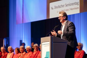 Der baden-württembergische Sozialminister Manne Lucha, ein Mann mittleren Alters, hält von einem Stehpult aus eine Rede. Er trägt einen dunklen Anzug und ein weißes Hemd. Neben ihm sitzen alten Menschen auf Stühlen, ein Chor. Sie alle tragen Kleidung in Rottönen. 