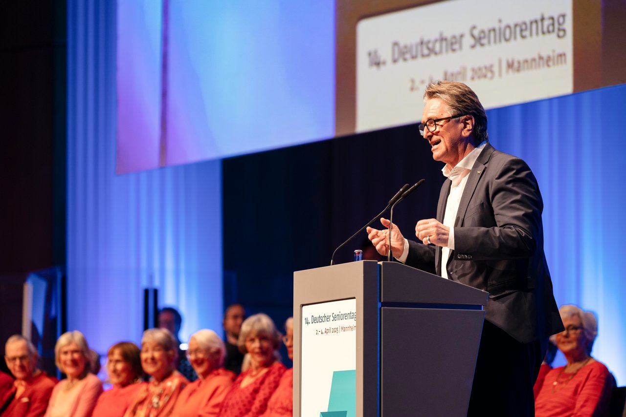 Der baden-württembergische Sozialminister Manne Lucha, ein Mann mittleren Alters, hält von einem Stehpult aus eine Rede. Er trägt einen dunklen Anzug und ein weißes Hemd. Neben ihm sitzen alten Menschen auf Stühlen, ein Chor. Sie alle tragen Kleidung in Rottönen.  Klick öffnet eine vergrößerte Ansicht.