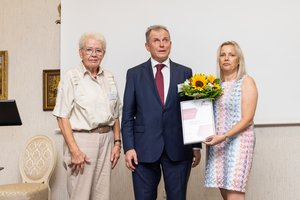 Zwei Frauen und ein Mann stehen auf der Bühne. Eine Frau hält einen Blumenstrauß und ein Zertifikat in der Hand.