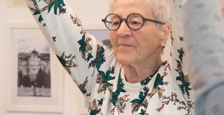 Ältere Frau mit Brille hebt ihre Hände über den Kopf. Sie schaut konzentriert.
