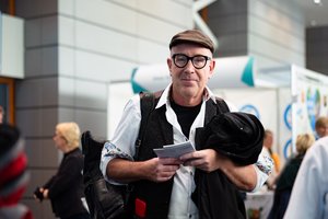 Ein älterer Mann mit Cord-Käppi auf dem Kopf und einer schwarz geränderten Brille. Er hat ein weißes Hemd an, darunter ein schwarzes T-Shirt und darüber eine dunkle Weste. Er hält eine Broschüre in der Hand und über dem Arm trägt er seine Jacke.