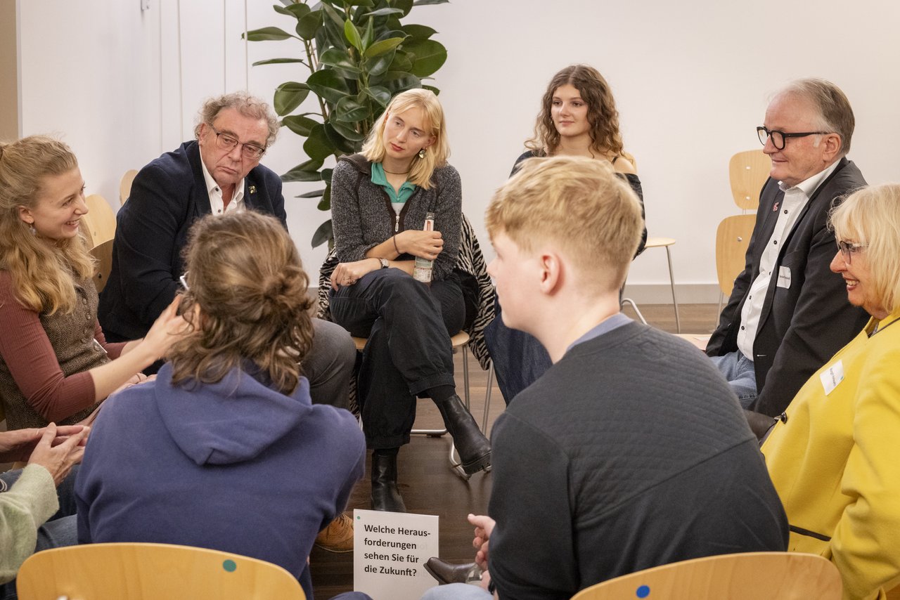 Eine Gruppe junger und älterer Menschen sitzen in einem Stuhlkreis zusammen. Sie unterhalten sich. Klick öffnet eine vergrößerte Ansicht.
