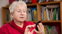 Ein alte Frau mit kurzen grauen Haaren und roten Pullover sitzt vor einem Bücherregal. Sie hat ein Smartphone in der Hand und schaut darauf.