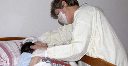 Sterbende im Pflegebett, der sich eine jüngere Frau in Schutzkleidung liebevoll zuwendet.
