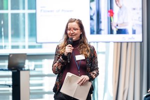 Eine junge Frau mit langem rotem Haar und Brille steht vor der Zuhörerschaft. Sie hat Zettel in der Hand und spricht in ein Mikrofon. Im Hintergrund sieht man einen großen Bildschirm.