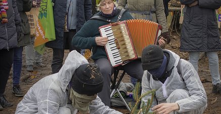 Zwei Menschen in weiß halten Tannenäste.Dahinter eine alte Frau, die Akkordeon spielt. Dahinter eine Menschenreihe