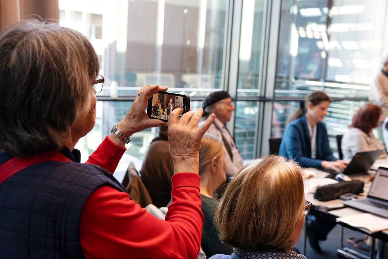 Eine ältere Frau hält ihr Smarphone zum Fotografieren in der Hand. Sie will eine Szene im Raum mitfilmen: An einem Tisch sitzen Menschen rund um einen Tisch auf dem Coputer und Laptobs stehen. In der Mitte sprecht ein Mann zu ihnen. Klick öffnet eine vergrößerte Ansicht.