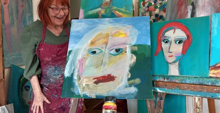Ältere Frau in einem Atelier zeigt farbenfrohe Portraits.