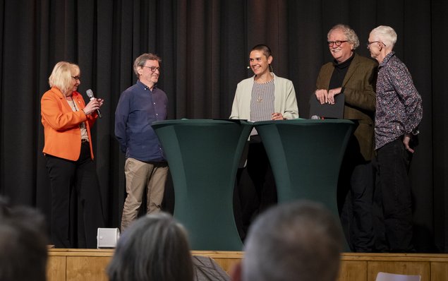 Auf der Bühne stehen zwei Stehtische. Um diese Tische steht eine kleine Gruppe. Eine Frau spricht gerade in ein Mikrofon. Klick öffnet eine vergrößerte Ansicht.