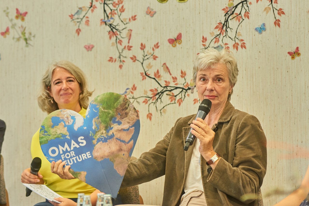 Zwei Frauen, die eine hält ein herzförmiges Plakat in der Hand auf dem steht "Omas for Future". Beide halten einen Mikrofon und schauen in die Kamera. 