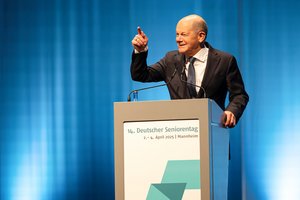 Der Bundeskanzler Olaf Scholz, ein älterer Mann mit Halbglatze, trägt einen dunklen Anzug, ein weißes Hemd und eine schwarz Kravatte. Er steht am Rednerpult. Er hebt seine rechte Hand mit gestrecktem Zeigefinger und lächelt. Der Hintergrund ist blau.