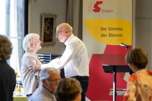 Ein älterer Mann und eine ältere Frau begrüßen sich herzlich. Die lächeln sich an und der Mann berührt die Frau am Oberarm.