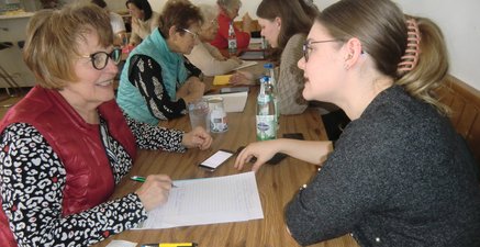 Seniorinnen sitzen an einem langen Tisch. Gegenüber sitzen Jugendliche, die den Senioren die Funktion des Handys erklären.