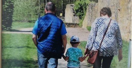 Zwischen einem älteren Paar (Mann und Frau) geht ein kleines Kind. Es fasst beide an der Hand.
