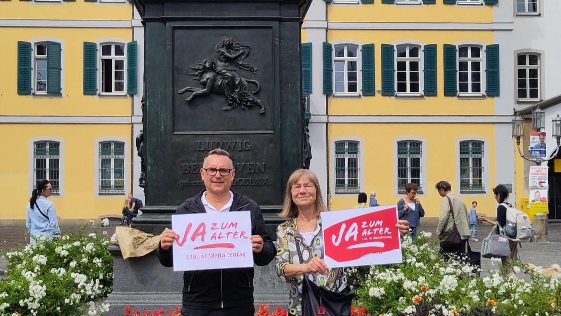 Ein Mann und eine Frau stehen vor einem Denkmal, von dem nur der Sockel zu sehen ist.  Sie halten beide jeweils ein Schild in der Hand mit der Aufschrift "Ja zum Alter!" Klick öffnet eine vergrößerte Ansicht.